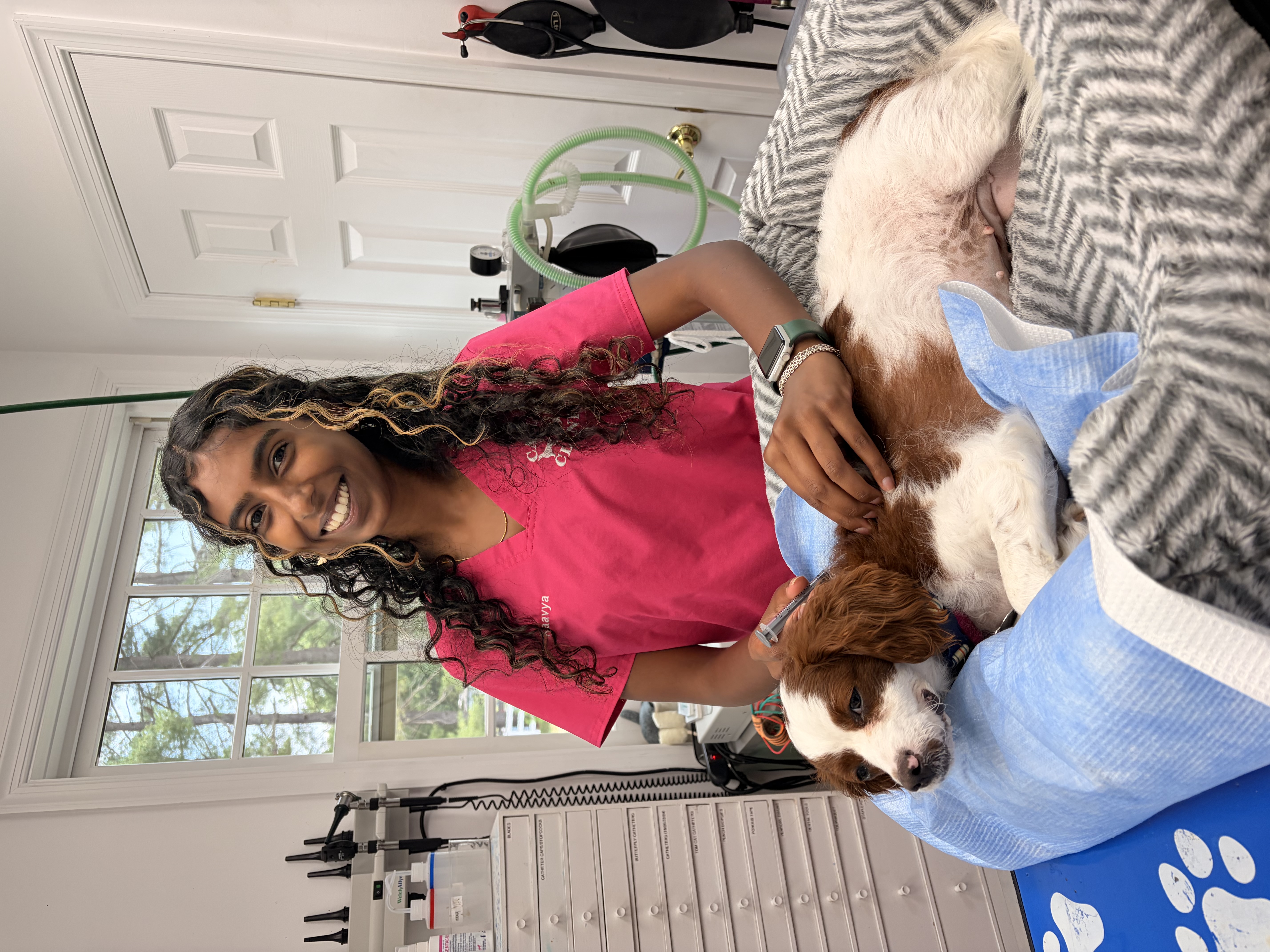 A woman with curly brown hair and blonde highlights wears pink scrubs and poses with a brown and white dog in a dog bed. Behind her are medical devices, tubing, and a window. 